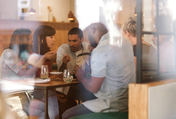 Gruppo di giovani amici diversi seduti a un tavolo all'interno di un bistrot alla moda che si godono un pasto insieme
 - Foto, immagini