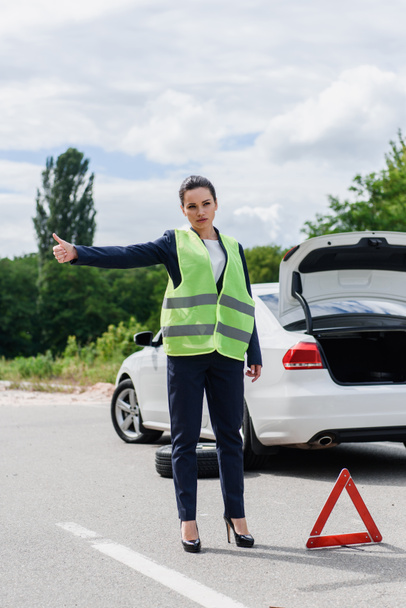 attractive businesswoman in light green vest hitchhiking on road near broken car - Photo, Image