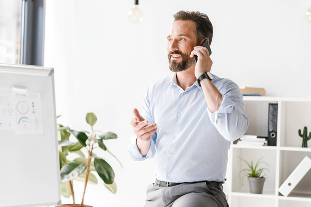 Smiling businessman talking on mobile phone while leaning on a office desk - Foto, Bild