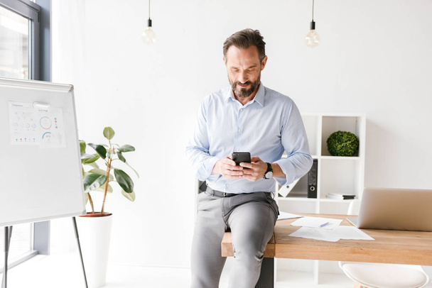 Smiling businessman using mobile phone while leaning on a office desk - Foto, Imagem