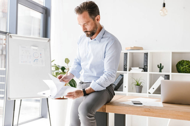 Focused businessman looking at paperwork while leaning on a office desk - Fotó, kép