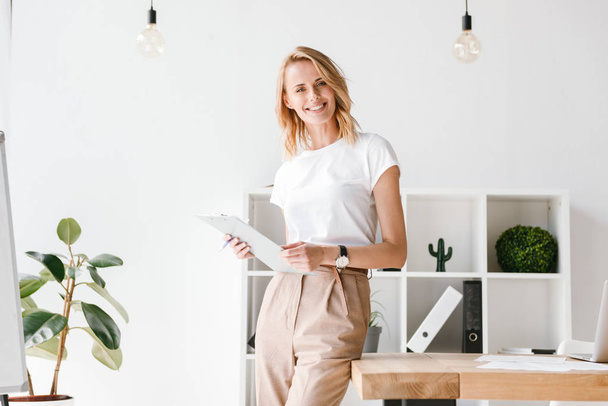 Smiling businesswoman holding folder while leaning on a office desk - Foto, imagen