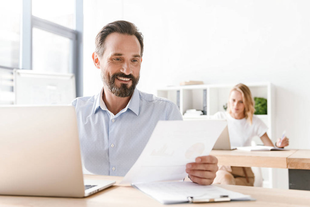 Hombre sonriente gerente que trabaja en el ordenador portátil y documentos mientras está sentado en la oficina con la mujer compañero de trabajo en un segundo plano
 - Foto, imagen
