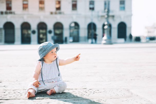 Baby girl sitting on a european city street and pointed finger up.  Little child outdoors in a town. Architecture of Trieste with pedestrian, people, Italy, Europe. - Foto, imagen