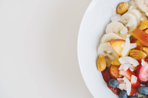 Λευκό πιάτο με βρώμη, μπανάνα, αμύγδαλο, μυρτιλός, ροδάκινο και καρύδα νιφάδες. Υγιεινό πρωινό. Ο Κόουπ χώρο, το top view - Φωτογραφία, εικόνα