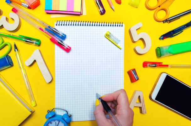 the student makes notes in a notebook. copy space. school accessories on a desk on a yellow background. concept of education. stationery. watches, colored pens, phone, markers. view from above. top view. flat lay - Photo, Image