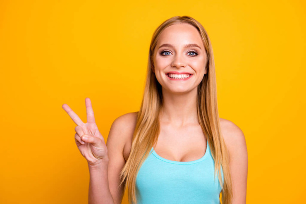 Portrait of young woman with blond hair, big blue eyes and toothy beaming smile. Positive girl demonstrating two fingers v-sign isolated on yellow background with copy space - Фото, изображение