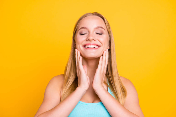 Young charming blonde girl closes her eyes with joy and touches her cheeks with her hands against yellow background - Foto, Bild