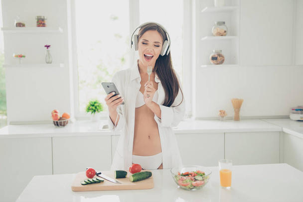 Good morning! Hot tempting and seductive woman in white lingerie and shirt with long sleeve, listening music through headphones and sings in the kitchen holds fork as a microphone - Fotografie, Obrázek