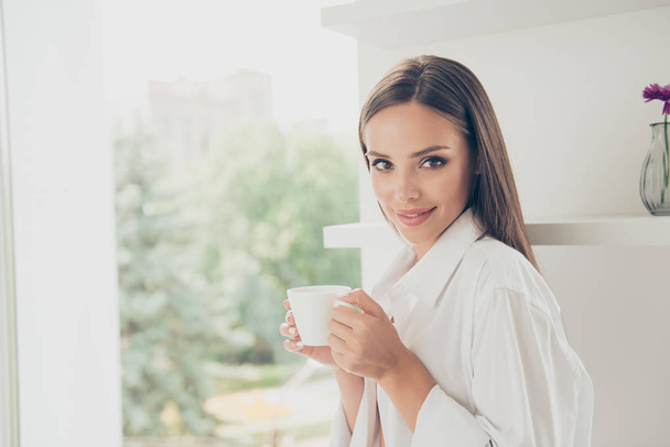 Ritratto ravvicinato di una giovane donna attraente in camicia bianca durante una pausa caffè mattutina, si riposa e si gode il drink, guarda la fotocamera e sorride
 - Foto, immagini