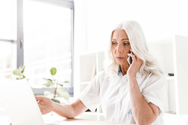Image of serious mature woman sitting indoors in office working talking by mobile phone using laptop computer. - Foto, afbeelding