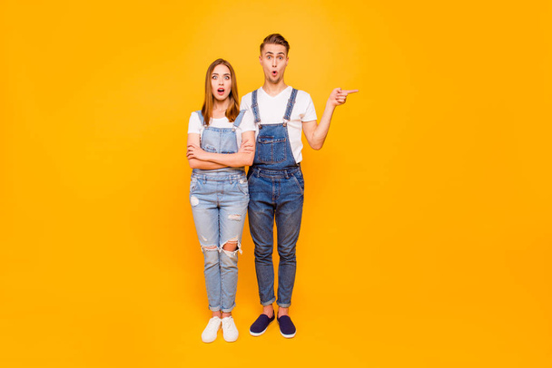Full lenght portrait of cute beautiful young couple, astonished with widely opened mouths over yellow background. Guy is pointing left with his finger, isolated, copyspace - Valokuva, kuva