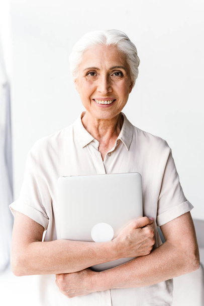 Happy mature business woman hugging laptop computer while standing at the office and looking at camera - Foto, Imagem