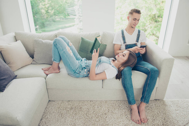 Young attractive beautiful cute couple wearing overall, home on sofa barefoot. Girl lying and reading book, guy surfing the Internet with his gadget - Foto, Bild