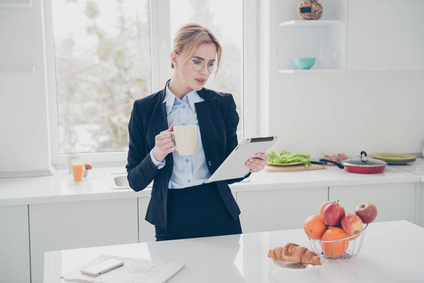 Adorable beautiful blonde woman office executive worker wearing stectacles early in the morning wearing dark blue jacket and skirt having a drink with croissant. Mobile phone lying on white table - Fotó, kép