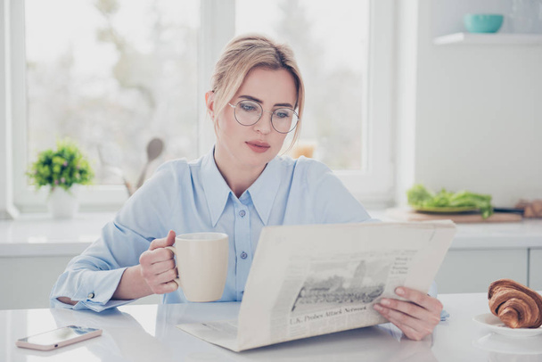 Adult adorable woman office executive worker wearing eye-glasses keeping in hands and reading newspaper early in the morning having a drink with croissant in light kitchen - Photo, Image