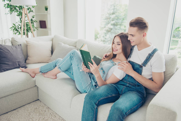 Young attractive adorable  cute couple wearing overall, home on sofa barefoot. Girl lying and reading book, guy hugging her shoulders - Fotó, kép