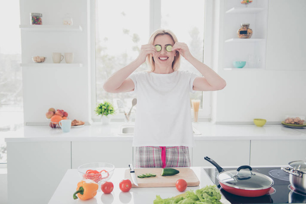 Attractive adorable beautiful charming nice smiling blonde woman wearing pajama putting chopped pieces of cucumber to her eyes having fun while cooking salad - 写真・画像