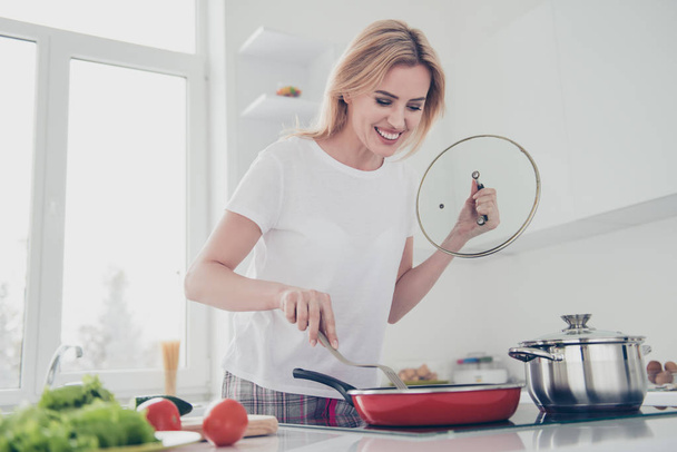 Adorable attractive adult smiling girl with manicure wearing pajama cooking stirring food on fry-pan with fork. Red tomatoes, green salad, cucumber lying on white table - Fotó, kép