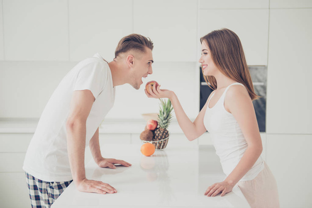 Portrait of young attractive cute lovely funky funny couple together in light fashionable kitchen, wearing pajama, playing, girl feeding guy with apple, man pretends eating fruit - Photo, Image