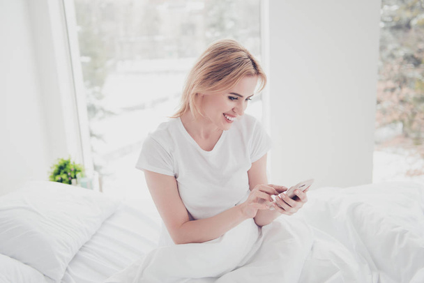 Young charming cute smiling blonde woman awake in bed on white sheet covered with blanket wearing t-shirt in bedroom with white interior texting sms on cell-phone. Big window on background - Valokuva, kuva