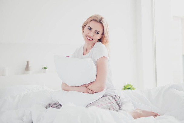 Joven linda mujer rubia sonriente despertando en la cama en sábanas blancas y manta abrazando almohada con pijama disfrutando en el dormitorio con interior blanco. Dieta
 - Foto, Imagen