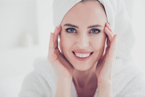 Close up portrait of young cute adult woman looking straight at camera smiling with her teeth wearing white bathrobe and turban keeping hands and fingers near eyes wrinkles in white interior - Foto, Bild