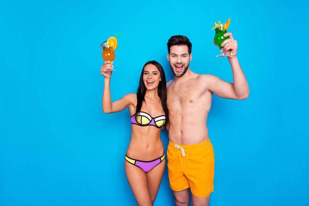 Beautiful young girl in swimsuit and handsome man in yellow shorts happily shout out a greeting and raise glasses with drinks up! Couple rest guys on blue background with place for text - Photo, Image