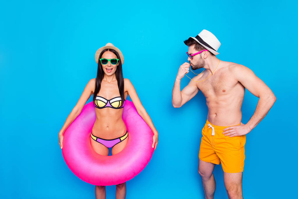 Muscular man in yellow shorts for swimming blows into the whistle and looks at the happy girl in a colorful swimsuit which holds an inflatable lifebuoy around her waist - Foto, afbeelding