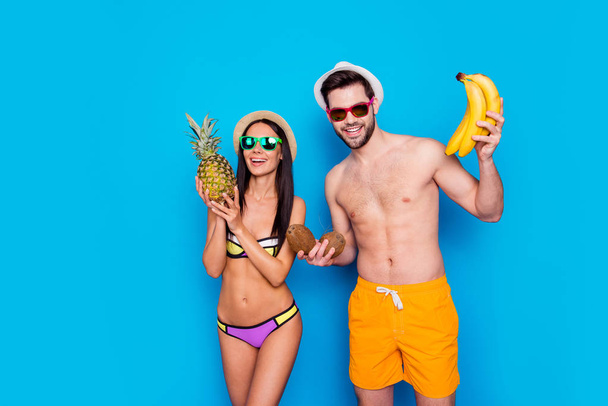 Fiatal és bájos hölgy öltözött fürdőruha tart egy ananász álló mellett egy férfi sárga nadrág, trópusi gyümölcsökkel, a kezét a kék háttér elszigetelt. Vitaminok és egészséges nyári koncepció - Fotó, kép