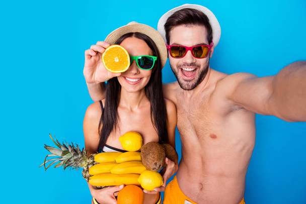 Concepto de vacaciones de verano. Chico y chica vestidos con ropa de playa sostienen frutas tropicales hacen selfie en la cámara frontal del teléfono inteligente de pie aislado sobre fondo azul
 - Foto, Imagen