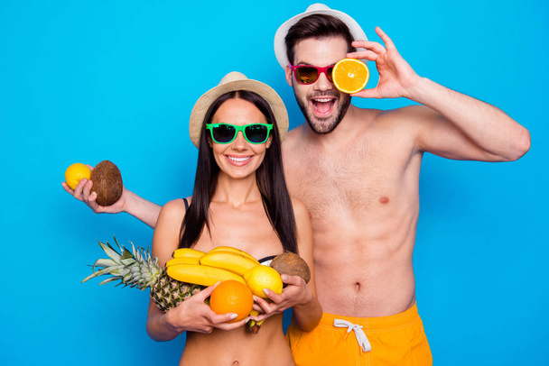 Handsome couple in in a good mood and colourful eyewear holding a lot of fruit. Funky young woman and handsome men messing around with fruit - Foto, afbeelding