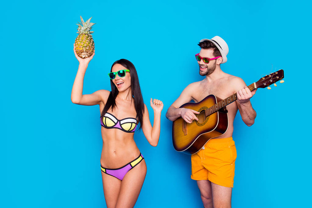 Time to celebrate vacations! Brunette guy with beard plays on guitar in hot summer day while a beautiful and charming girl dances to his music with pineapple in her hand isolated on blue background - Photo, image