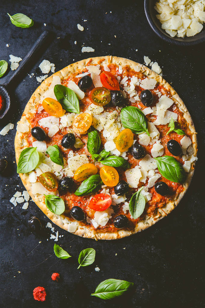 Top view of tasty appetizing classic italian traditional pizza with cheese, olives, basil and fresh vegetables on dark black table.  - Фото, зображення