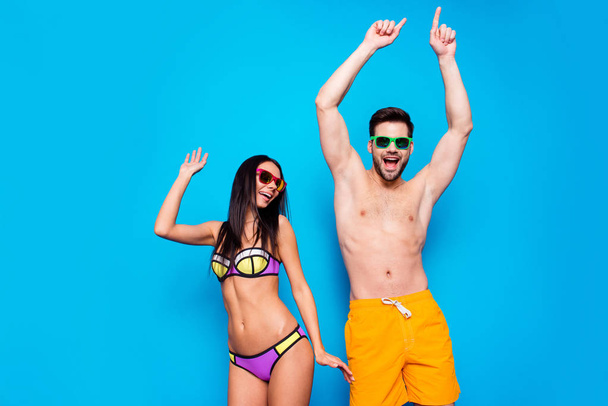 Hot summer time concept. Portrait of cheerful couple in sunglasses and bright swimsuits dance together with beach party mood raise their hands up isolated on bright blue background - Photo, Image