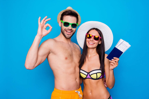 Ready to journey or burning tour! Close up portrait of charming, pretty couple in glasses with passport and flying tickets in hand, gesture ok sign, isolated on blue background - Foto, Imagen