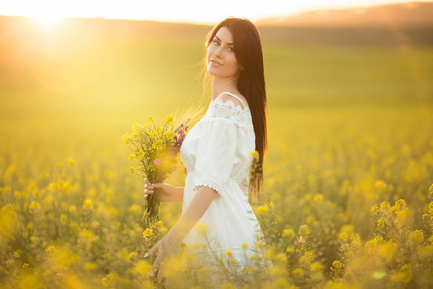 Happy young woman with bouquet of wildflowers in yellow field in sunset lights - Foto, afbeelding