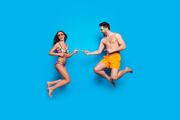 Full length portrait of a joyful young woman and happy man in eyewear, dressed in swimsuit, jumping and show fingers on each other over blue background with copy space for text - Fotoğraf, Görsel