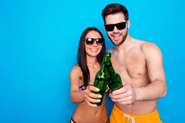 Handsome guy and young woman dressed in glasses, colored swimsuits, holding cold drink in glass bottle and celebrate beginning of holiday isolated on blue background with copy space for text - Foto, afbeelding