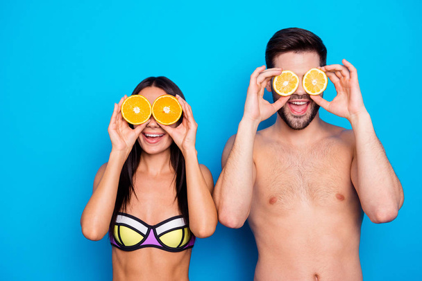 Cheerful young woman ang attractive muscles guy holding two halves of orange and lemons near eyes isolated on blue background. Healthy summer concept - Foto, afbeelding