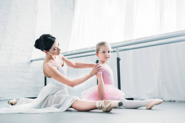 очаровательная маленькая балерина в розовой пачке сидит и растягивается во время тренировки с учителем балета в студии
 - Фото, изображение