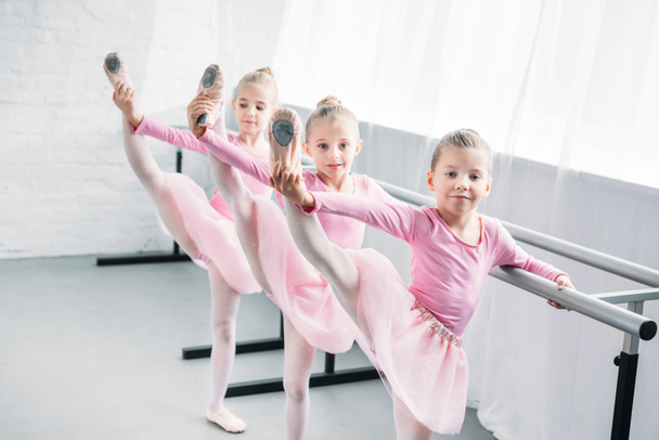 adorable kids in pink tutu skirts practicing ballet and looking at camera in ballet school - Zdjęcie, obraz
