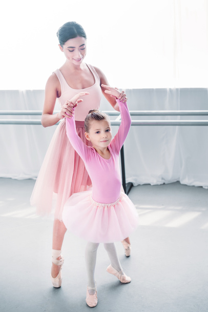 adorable child in pink tutu looking at camera while training with teacher in ballet school - Photo, Image