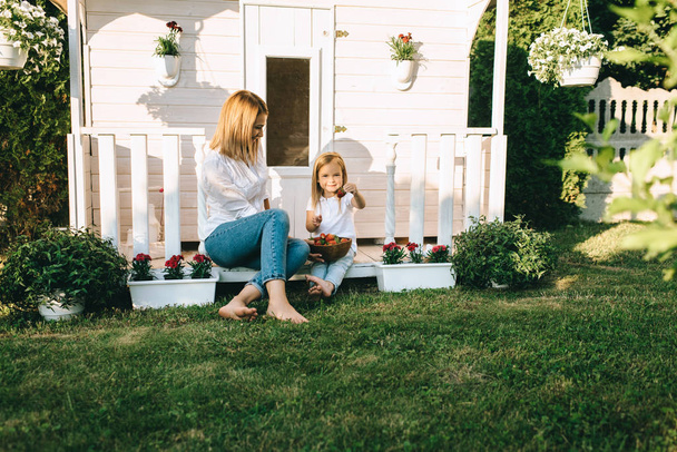 Madre y adorable niño pequeño con tazón de fresas descansando en el porche de la casa de campo juntos
 - Foto, imagen
