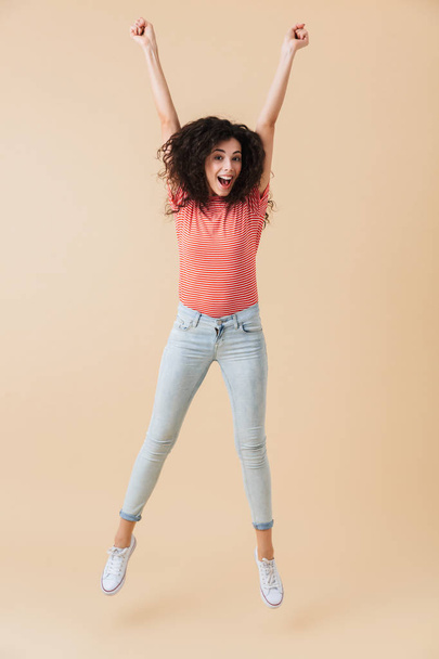 Image of pretty young woman jumping isolated over beige wall background looking camera. - 写真・画像