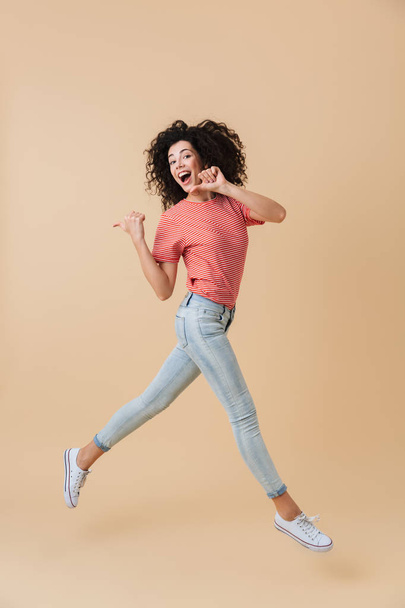 Full length portrait of a happy young woman with brunette curly hair jumping and pointing fingers away isolated over beige background - Φωτογραφία, εικόνα