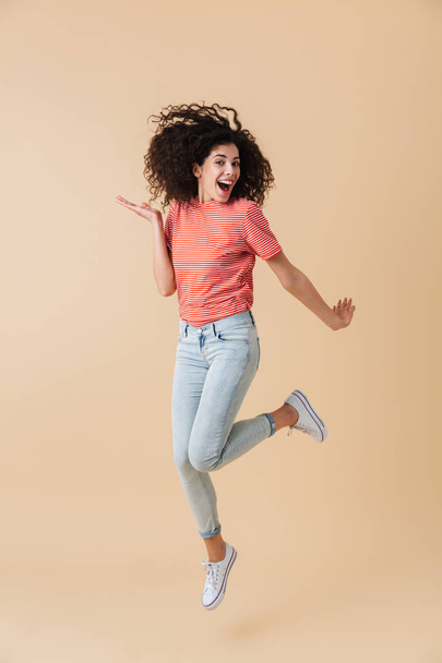 Full length portrait of an excited young woman with brunette curly hair celebrating while jumping isolated over beige background - Foto, Imagem