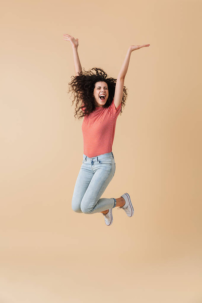 Full length portrait of a joyful young woman with brunette curly hair celebrating while jumping isolated over beige background - Fotografie, Obrázek