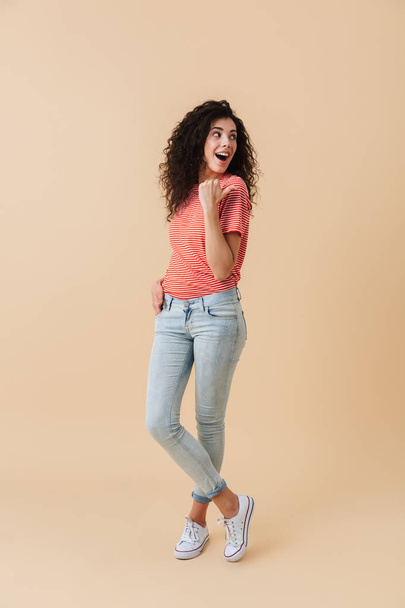 Full length portrait of a cheerful young woman with brunette curly hair pointing at copy space isolated over beige background - Fotoğraf, Görsel