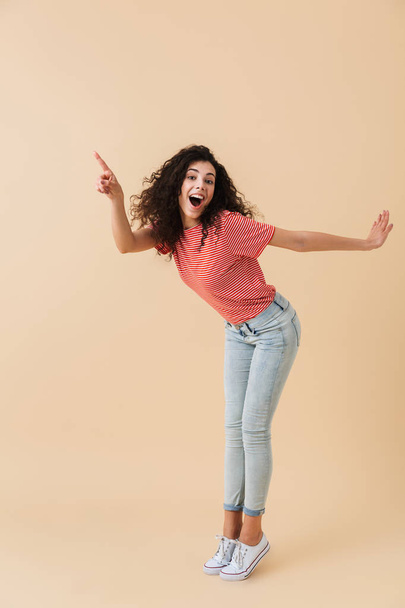 Full length portrait of a joyful young woman with brunette curly hair pointing at copy space isolated over beige background - Fotó, kép